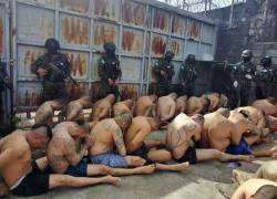 Fotografía de varios reclusos sentados ante filas de militares. Las imágenes recuerdan al control carcelario impuesto recientemente por El Salvador.