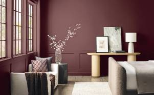 Habitación sofisticada con paredes en un profundo tono rojo marrón, molduras clásicas inferiores, un consola de madera clara de diseño minimalista y un sillón auxiliar con cojines decorativos.
