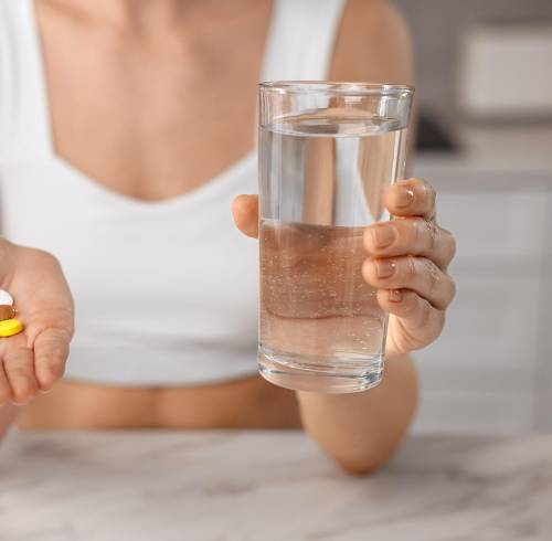 Mujer con un vaso de agua y pastillas en una mesa en el interior.