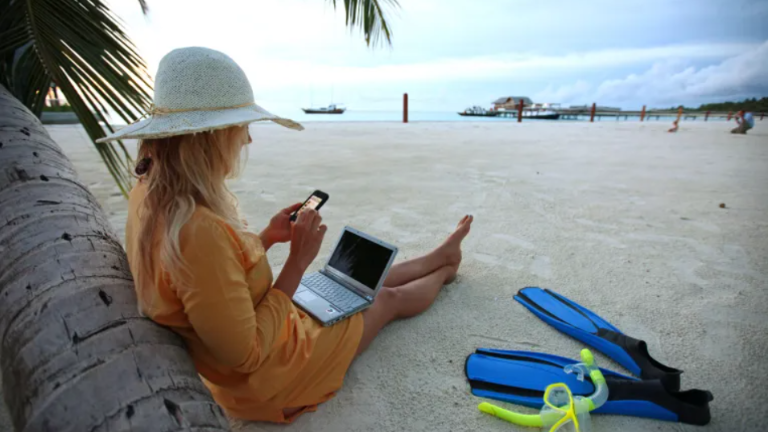 Una persona en la playa revisa su computador y atiende llamadas telefónicas.