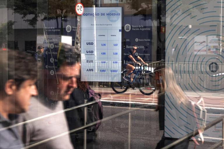 $!Personas caminan frente a un monitor que muestra el mercado de divisas este jueves, frente al Banco de la Nación en São Paulo.