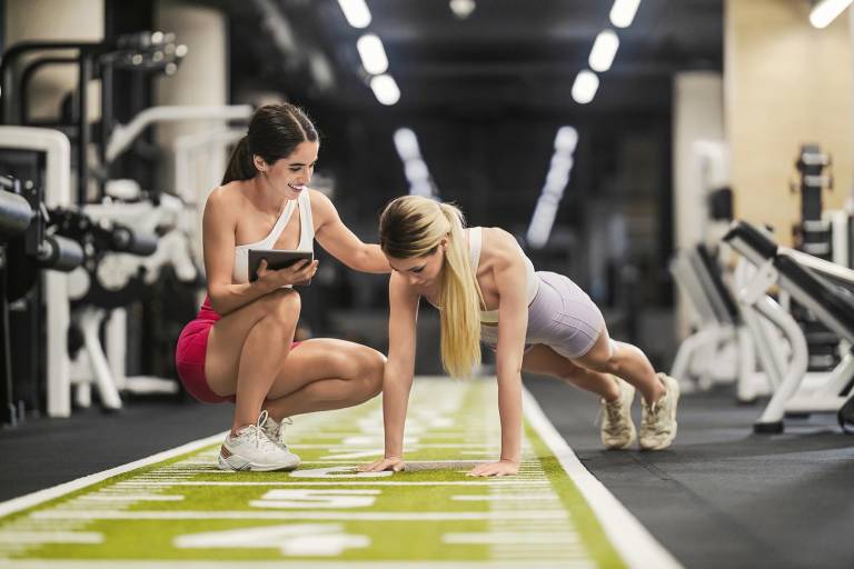 $!Atleta realiza flexiones en un gimnasio mientras su entrenador registra su progreso en una tableta.