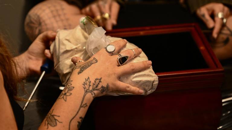 La tatuadora Kat Dukes (izquierda) abre una caja con las cenizas de la madre cremada de Scout Frank (derecha) para mezclarlas con la tinta de un tatuaje hecho a mano en Oceanside, California, el 6 de octubre de 2023.