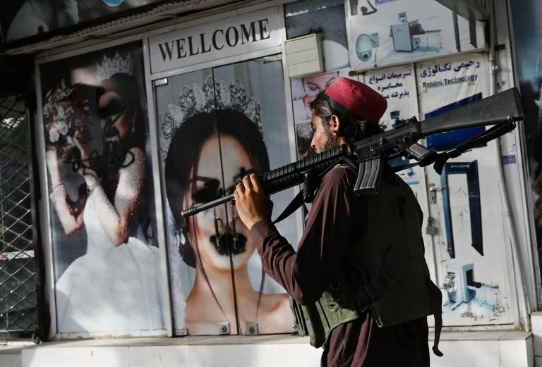 $!Un combatiente talibán pasa frente a un salón de belleza con imágenes de mujeres desfiguradas con pintura en aerosol en Shar-e-Naw en Kabul.
