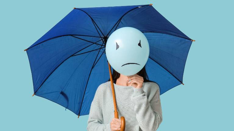 Mujer sosteniendo un paraguas azul y un globo con cara triste. Esta imagen representa el Blue Monday, el que se afirma es el día más triste del año.