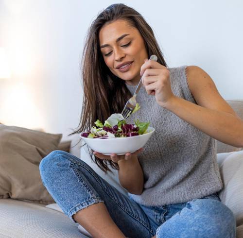 Mujer comiendo un bowl de ensalada en sofá.