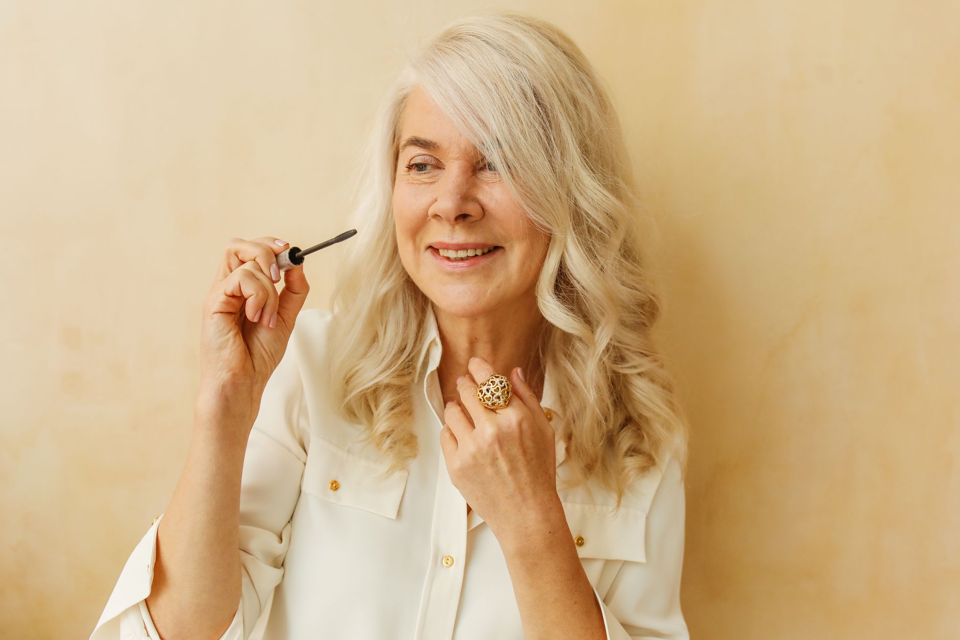 Mulher idosa com cabelo branco sorrindo enquanto se maquiva em ambientes internos.