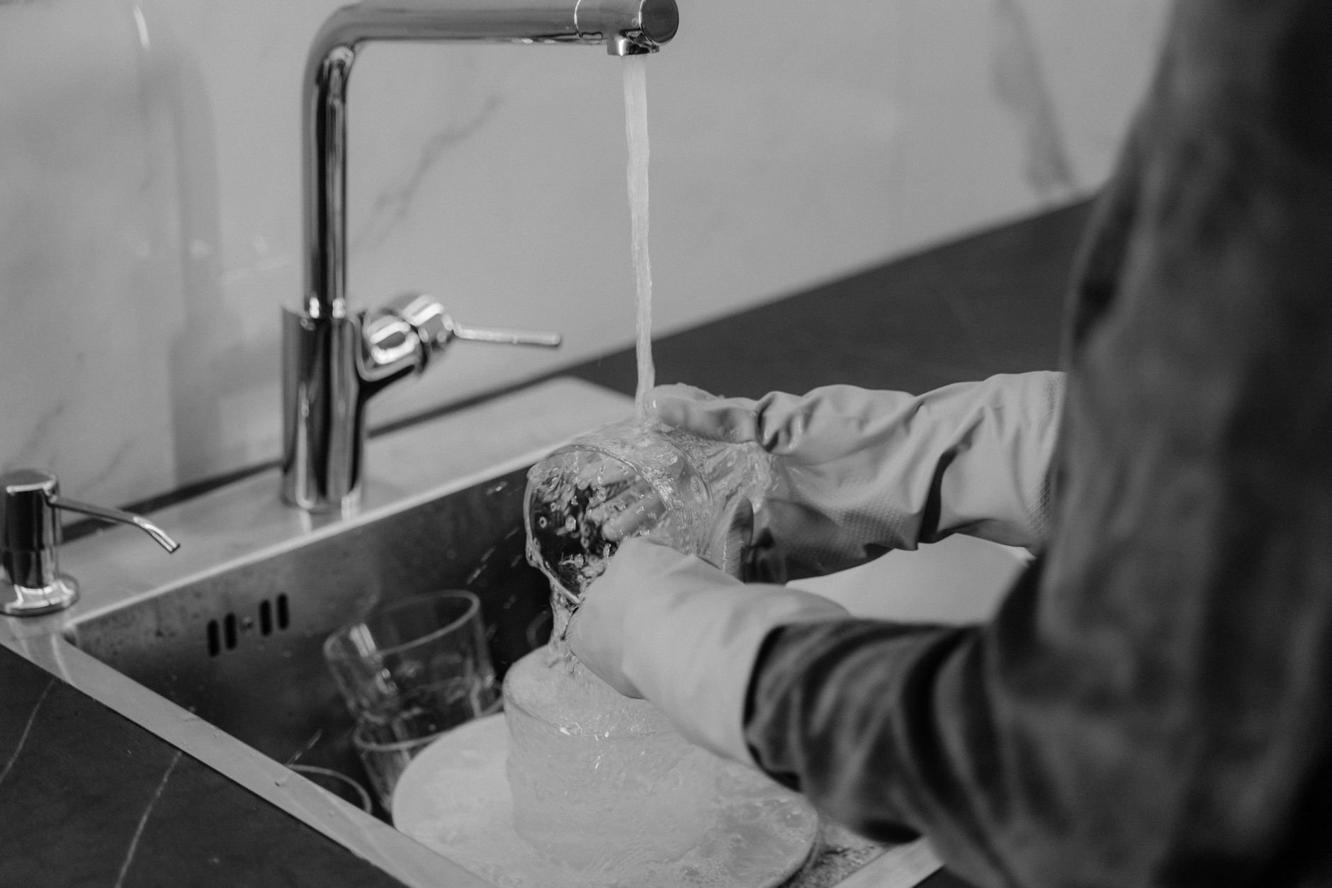 Foto em monocromático de uma pessoa lavando louça sob água corrente na pia da cozinha.